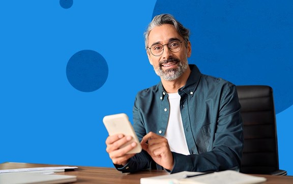 Uomo brizzolato, vestito in modo casual, alla scrivania con portatile e smartphone in mano.